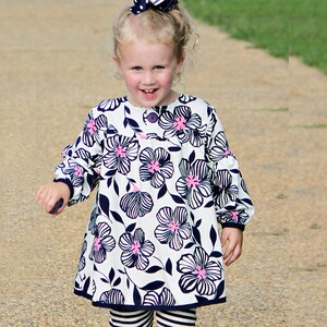 May include: A young girl wearing a white dress with a black and pink floral pattern. The dress has long sleeves and a button closure at the neck. The girl is smiling and looking at the camera.
