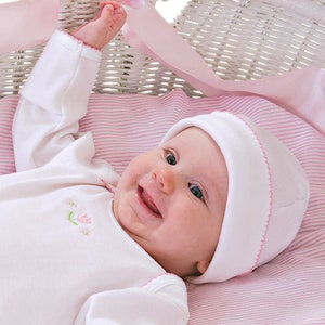 May include: A baby wearing a white long-sleeved onesie with pink embroidered flowers and a white hat with a pink trim. The baby is lying in a white wicker basket with a pink and white striped blanket.