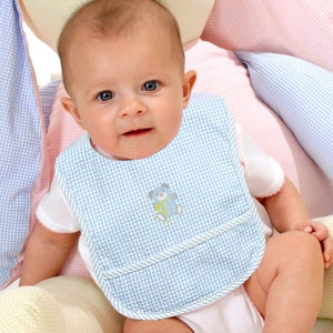 May include: A baby wearing a blue and white gingham bib with a small embroidered bear design. The bib has a pocket on the front.