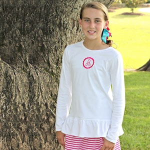 May include: White long-sleeved shirt with a pink ruffled hem and a pink circle with the letter "A" embroidered on the front.