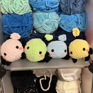 May include: A collection of colorful yarn balls in shades of blue and green, with four crocheted round creatures in pink, green, blue, and yellow. Each creature has black eyes, pink cheeks, and black feet. The yarn and creatures are in a clear plastic container.