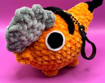 May include: A plush keychain shaped like a chainsaw character with a bright orange body, a textured gray blade, and a black handle. The character features a large circular black eye with a white border and includes a black plastic lobster clasp.