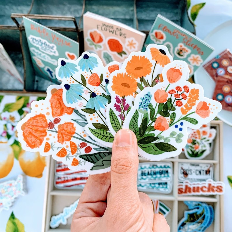 May include: A set of colorful floral stickers featuring blue, orange, and red flowers with green leaves. The stickers are arranged in a fan shape and are held in a hand.