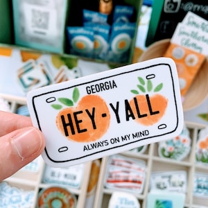 May include: A white sticker with a peach design and the text "Hey-Y'all" in black. The sticker is shaped like a license plate and has the words "Georgia" and "Always on my mind" in black text.