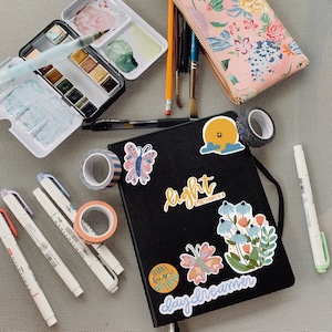 May include: A black journal with colorful stickers on the cover. The stickers include a sun, flowers, butterflies, and text that says "light", "daydreamer", and "shake loose, nothing's in your hands".