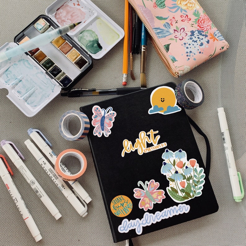 May include: A black journal with colorful stickers on the cover. The stickers include a sun, flowers, butterflies, and text that says "light", "daydreamer", and "shake loose, nothing's in your hands".