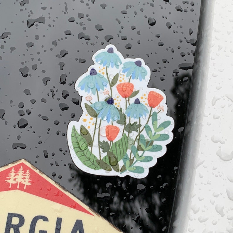 May include: A sticker featuring a cluster of blue and white flowers with orange centers, surrounded by green leaves. The sticker is on a black surface with water droplets.