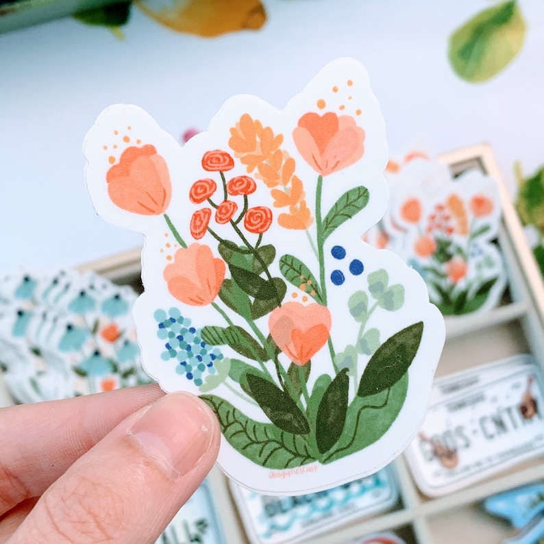 May include: A sticker featuring a bouquet of pink and orange flowers with green leaves and blue berries. The flowers are arranged in a cluster and have a whimsical, hand-drawn style.