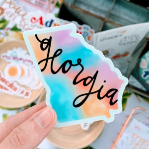 May include: A sticker featuring the outline of the state of Georgia with a watercolour-style background in shades of blue, pink, and orange. The word "Georgia" is written in black cursive script across the state outline.