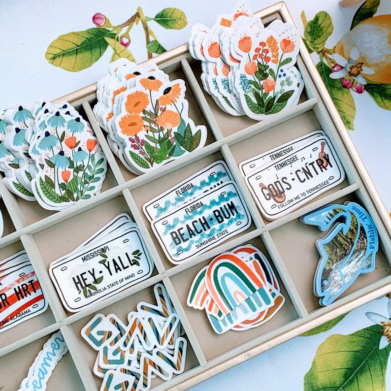 May include: A wooden tray filled with colorful stickers featuring various designs. The stickers include floral patterns, state-themed license plates, and a rainbow design. The tray is lined with a light brown fabric.