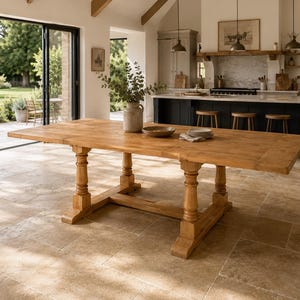 May include: A large rectangular wooden dining table featuring a natural light brown finish, a smooth flat top, and a sturdy base with four turned pedestal legs connected by a central horizontal support beam, positioned in a spacious room.