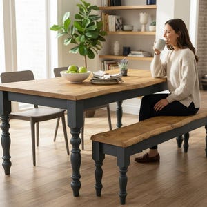 May include: A wooden dining table with a natural wood top and dark grey legs. A matching bench sits beside the table. A person is seated at the table. The room has a neutral colour palette with a large window and a plant.