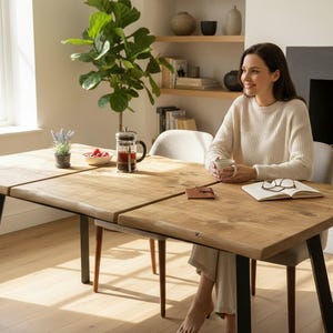 May include: A light brown wooden dining table with black metal legs. A woman sits at the table with a cup of coffee, a book, and glasses. A French press, a bowl of berries, and a potted plant are on the table.