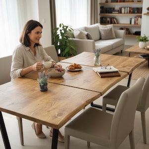 May include: A light brown wooden dining table with black metal legs. The table is set with a bowl of fruit, croissants, and a cup of coffee. Two chairs are positioned at the table. A woman is sitting at the table.