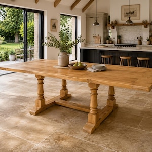 May include: A large rectangular wooden dining table with a natural light brown finish, featuring a thick tabletop and four sturdy turned pedestal legs connected by a central horizontal support beam, providing a stable base for the expansive surface.