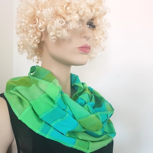 Op de afbeelding: Een groene en turquoise oneindige sjaal gemaakt van stofresten. De sjaal is gevouwen en gedrapeerd om de nek van een mannequin.