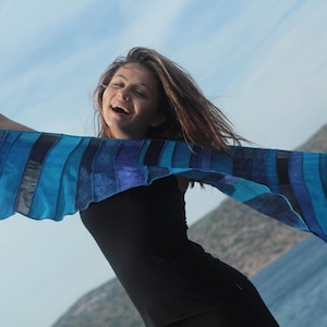 Op de afbeelding: Een vrouw draagt een zwart tanktop en een blauw-paarse gestreepte sjaal, glimlacht en houdt de sjaal voor zich.