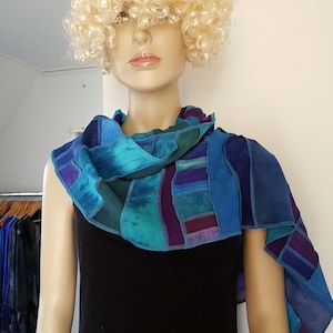 Op de afbeelding: Een rechthoekige sjaal met een patchworkontwerp van diverse blauwe, groenblauwe en paarse stofsegmenten. De sjaal is gedrapeerd rond de nek en schouders en toont een mix van effen en gemêleerde texturen in koele tinten met zichtbare stiksels.
