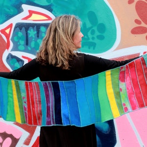 Puede incluir: Una bufanda de rayas coloridas hecha de retazos de tela. La bufanda es un arcoíris de colores, incluyendo rojo, naranja, amarillo, verde, azul, añil y violeta. La bufanda se está sosteniendo frente a una pared con grafitis.
