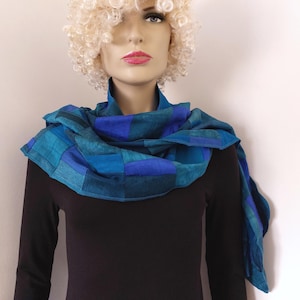 Op de afbeelding: Een blauwe en turquoise gestreepte sjaal met een geometrisch ontwerp. De sjaal wordt gedragen door een mannequin met blond haar.