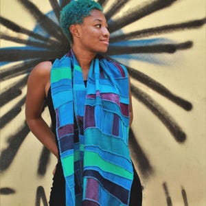 Könnte beinhalten: Eine Person mit blaugrünem Haar trägt einen blauen, grünen und violetten Patchwork-Schal. Der Schal besteht aus verschiedenen Stoffen und hat ein geometrisches Muster.