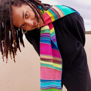 May include: A person with long dreadlocks wearing a black jacket and a colourful striped scarf. The scarf is tied around the neck and hangs down the front of the jacket.