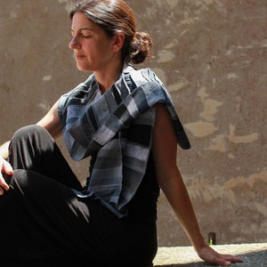 Op de afbeelding: Een patchwork sjaal in grijstinten en zwart is over de schouders gedrapeerd. De sjaal lijkt gemaakt van rechthoekige stukken stof die aan elkaar zijn genaaid. De persoon draagt een zwarte broek en een zwarte top, met gesloten ogen.