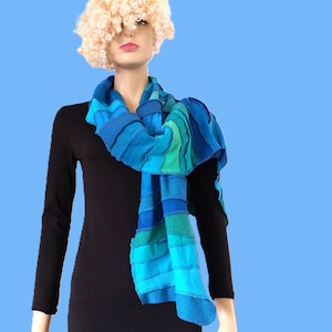 May include: A long scarf featuring horizontal stripes in various shades of blue, teal, and green. The scarf is constructed from multiple fabric panels with visible stitching, creating a textured, layered appearance draped around the neck.