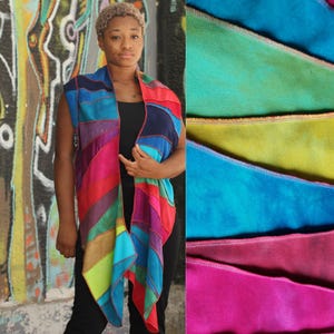 Può includere: Una sciarpa patchwork colorata nei toni del rosso, blu, viola e verde. La sciarpa è indossata sopra un top nero. L'immagine mostra anche un primo piano del tessuto in vari colori. Lo sfondo presenta graffiti.