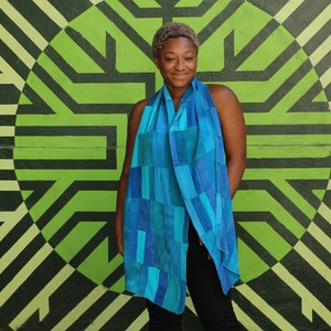 Op de afbeelding: Blauwe sjaal met een patchwork ontwerp in tinten turquoise en blauw. De sjaal is over de schouders gedrapeerd. De achtergrond is een groen en zwart geometrisch muurschildering.