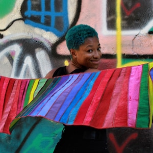 May include: A person with teal blue hair is holding a colourful striped fabric scarf in front of a graffiti wall.