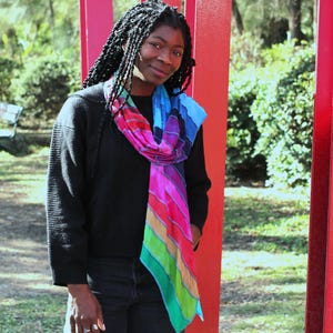 May include: A person wearing a black jumper and jeans, with a vibrant rainbow scarf. The scarf features stripes of blue, purple, pink, red, green, and yellow. The background includes red pillars and greenery.