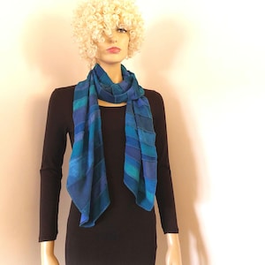 Op de afbeelding: Een lange sjaal met een verloop van blauwe en turquoise horizontale strepen. De sjaal is om de nek gedrapeerd en hangt aan de voorzijde naar beneden, met een verscheidenheid aan tinten variërend van diep marineblauw tot helder turquoise.