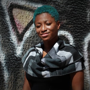 Op de afbeelding: Een persoon met tealblauw haar draagt een zwart-grijze gestreepte sjaal. De sjaal is gemaakt van stof dat gerecycled of upcycled lijkt te zijn. De persoon staat voor een muur met graffiti.