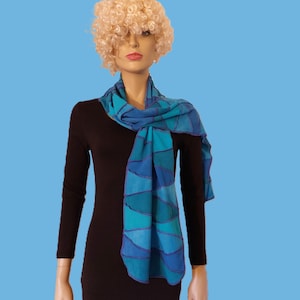 May include: A blue and teal scarf with a geometric pattern is draped around a mannequin's neck. The scarf features a series of curved shapes in varying shades of blue and teal, outlined with darker blue lines. The mannequin is wearing a black dress.