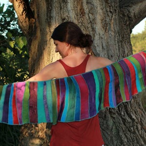 Op de afbeelding: Een vrouw in een rood tanktop houdt een kleurrijke gestreepte sjaal vast die is gemaakt van aan elkaar genaaide stofstroken. De sjaal is gemaakt van verschillende tinten groen, blauw, paars en rood.