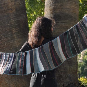 Könnte beinhalten: Eine Frau trägt einen langen, gestreiften Schal in verschiedenen Blautönen, Grau- und Weißtönen. Der Schal ist über ihre Schultern gelegt und wird in ihrer rechten Hand gehalten. Sie steht vor einem Baumstamm.