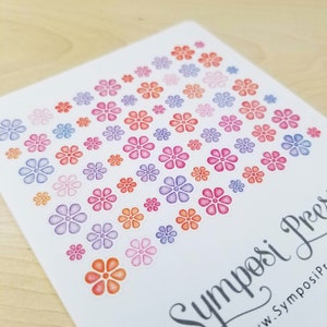May include: A sheet of colorful flower stickers in shades of pink, orange, purple, and blue. The stickers are arranged in a gradient pattern, with the darkest colors at the top and the lightest colors at the bottom. The sheet is labeled "Symposi Press" at the bottom.