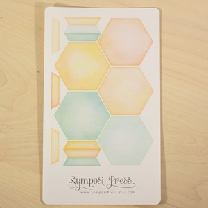 May include: A white card featuring watercolor-style geometric shapes in pastel colors: peach, yellow, and teal. The design includes hexagons and rectangular shapes. The text "Symposi Press" and a website address are at the bottom.