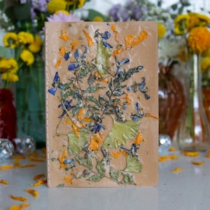 Op de afbeelding: Een bruine journalhoes met geperste bloemen en bladeren. De bloemen zijn blauw, geel en groen. Het journal ligt op een tafel met verspreide gele bloemblaadjes.