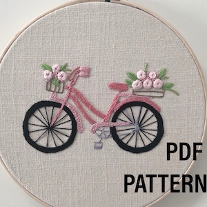 Puede incluir: Diseño de bicicleta bordada en rosa con ruedas negras y una cesta de flores rosas. El bordado está colocado en un aro de madera redondo sobre una tela beige claro. El texto en la imagen dice "PDF PATTERN".
