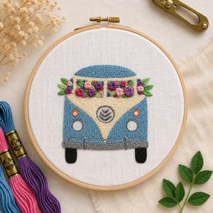 Puede incluir: Arte de aro bordado con una furgoneta vintage azul y crema con una guirnalda floral. La furgoneta tiene neumáticos negros y molduras grises. El bordado está en un aro de madera. Hilo de bordar colorido visible en la esquina.