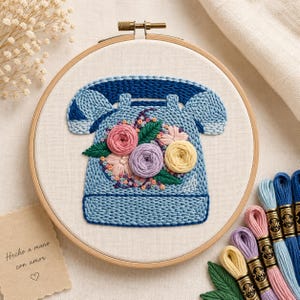 Puede incluir: Arte de aro bordado con un diseño de teléfono azul claro con un arreglo floral de flores rosas, moradas y amarillas. El bordado está en un aro de madera. También se incluye hilo de bordar y una pequeña tarjeta con texto.