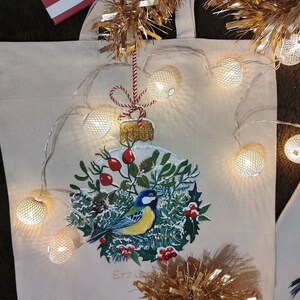 May include: A white canvas tote bag with a painted Christmas ornament featuring a bluebird and red berries. The ornament is surrounded by a garland of green leaves and white snow. The bag is decorated with a string of white lights and gold tinsel.