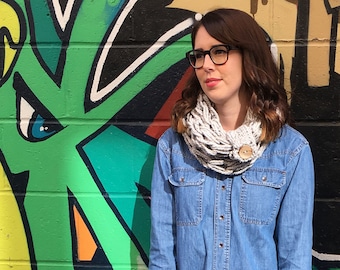 May include: A woman wearing a light blue denim shirt, a white knit scarf, black leather leggings, and black Chelsea boots. She is standing in front of a colorful graffiti wall.