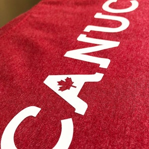 May include: Red t-shirt with white text that reads "CANUCK" with a Canadian maple leaf in the "A".