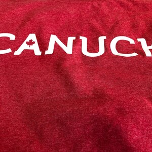 May include: Red t-shirt with white lettering that says "CANUCK" with a small Canadian maple leaf in the "A".