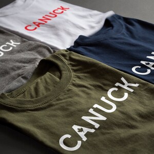 May include: Four folded t-shirts with the word "CANUCK" printed on them. The shirts are in different colors: gray, white, navy blue, and olive green. The word "CANUCK" is printed in white on the gray, navy blue, and olive green shirts. The word "CANUCK" is printed in red on the white shirt.