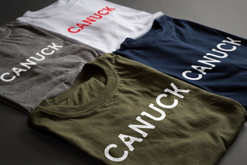 May include: Four folded t-shirts with the word "CANUCK" printed on them. The shirts are in different colors: gray, white, navy blue, and olive green. The word "CANUCK" is printed in white on the gray, navy blue, and olive green shirts. The word "CANUCK" is printed in red on the white shirt.