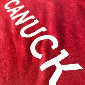 May include: Red t-shirt with the word "CANUCK" printed in white on the front.